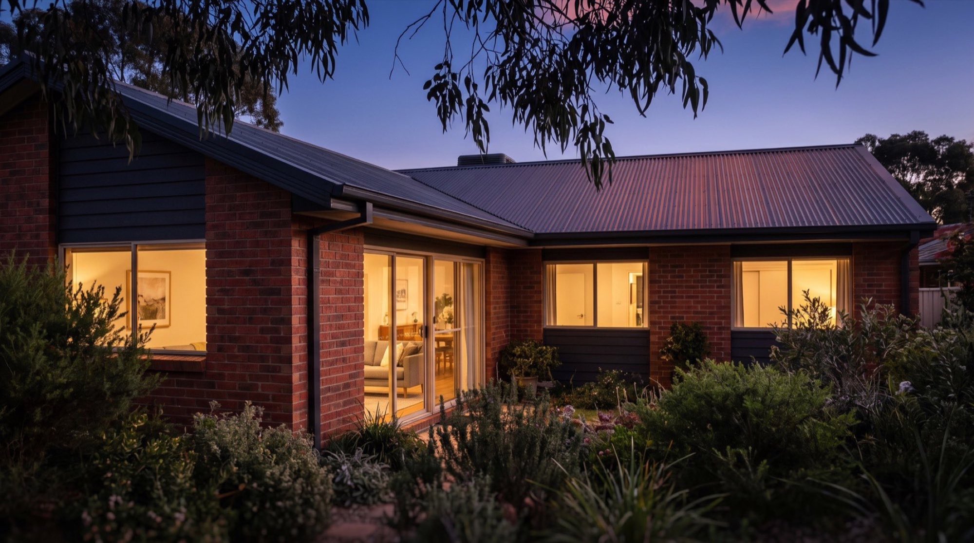 A brick home with lit windows at dusk under a gum tree