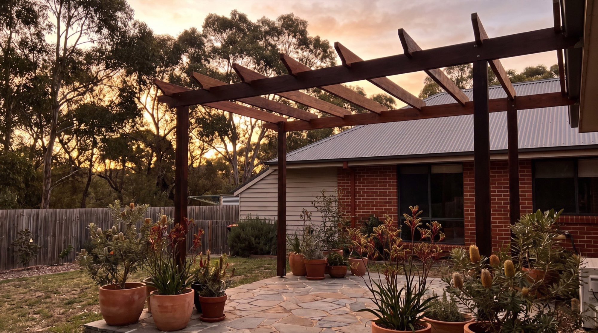A timber pergola over a paved patio beside a brick house at sunset